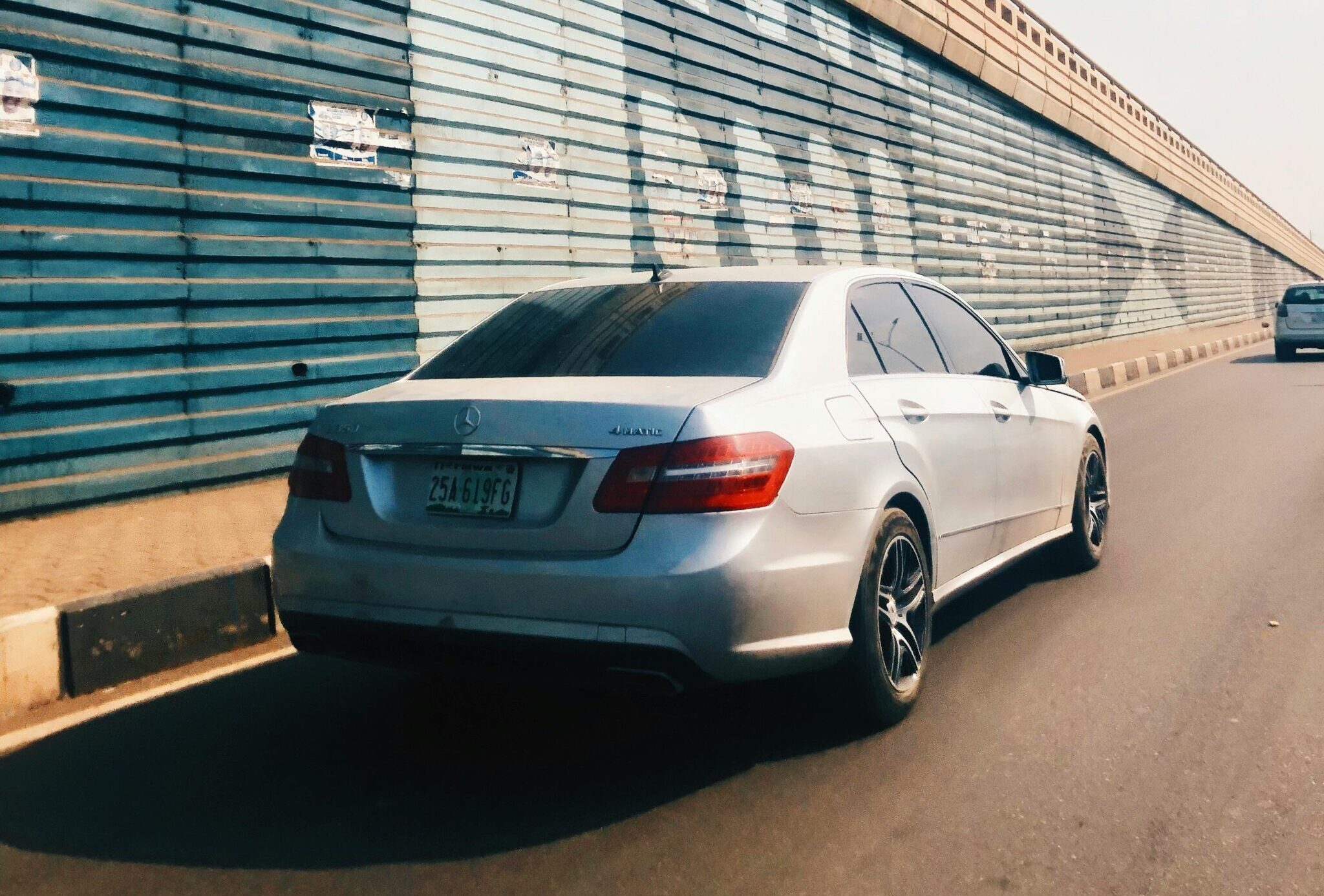 A silver car drives along a city street lined with a blue painted wall on a sunny day.